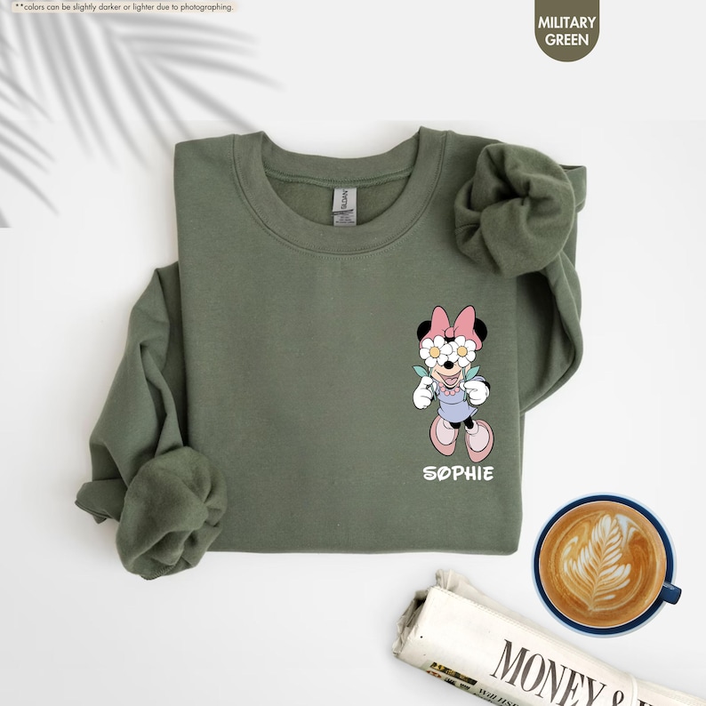 May include: Military green sweatshirt featuring a cartoon Minnie Mouse design with a floral headband and the name "Sophie" in white. The sweatshirt has long sleeves and a crew neck. A cup of coffee and a newspaper are in the background.