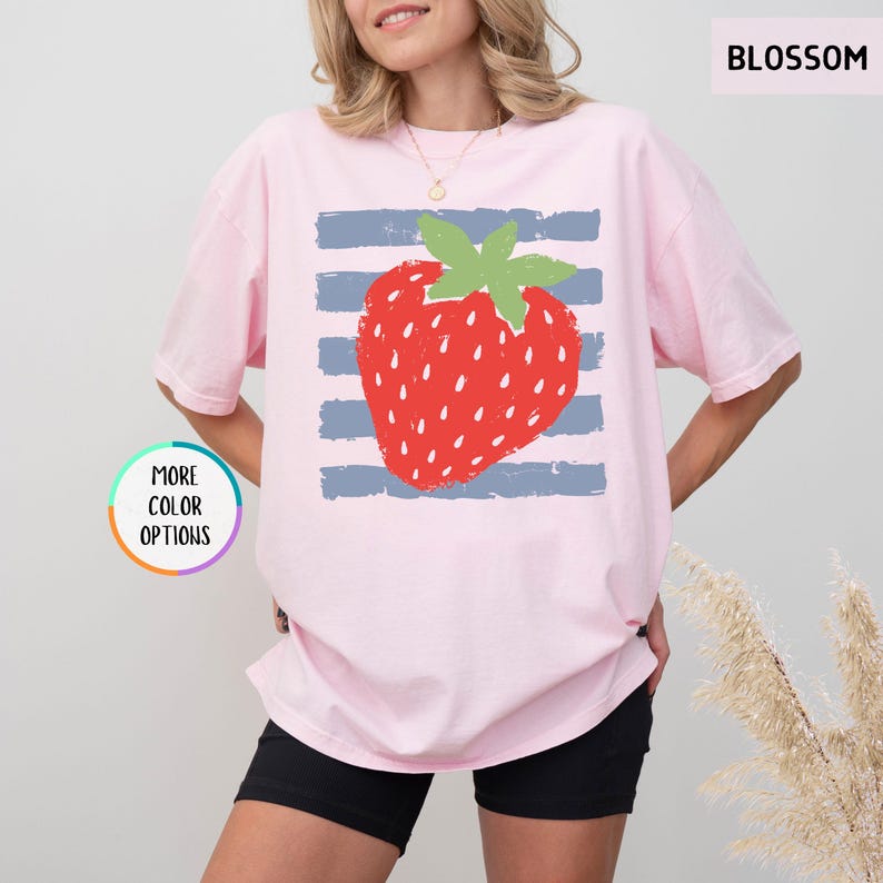 May include: A light pink short-sleeve t-shirt featuring a graphic of a red strawberry with a green stem centered over horizontal blue stripes. The text BLOSSOM appears in the top right corner, and a circular badge reads MORE COLOR OPTIONS.
