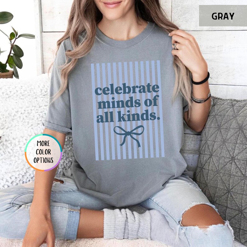 Puede incluir: Camiseta gris con el texto azul "celebrate minds of all kinds", sobre un fondo a rayas azul claro y un lazo azul. La camiseta es de estilo casual, ideal para el uso diario.