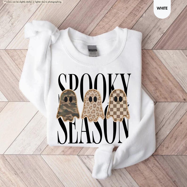 May include: A white long-sleeve sweatshirt featuring the black text SPOOKY SEASON and three ghost illustrations. The ghosts have different patterns: one with camouflage, one with a leopard print, and one with a brown and white checkerboard pattern.