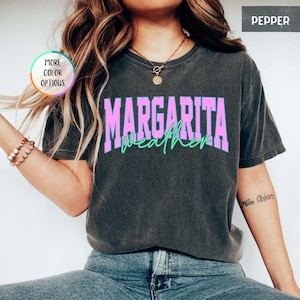 May include: A dark gray t-shirt with the words "MARGARITA weather" in a pink and green design. The shirt has a crew neck and short sleeves. The shirt is made of a soft, comfortable material.