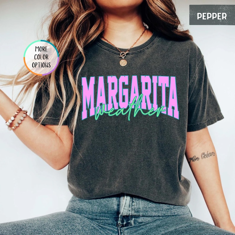May include: A dark gray t-shirt with the words "MARGARITA weather" in a pink and green design. The shirt has a crew neck and short sleeves. The shirt is made of a soft, comfortable material.