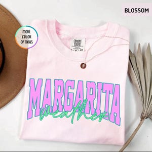May include: A light pink t-shirt with the words "MARGARITA WEATHER" in large, bold, pink and blue letters. The shirt is made of a soft material and has a round neckline. A gold necklace with a pendant is draped on the shirt.