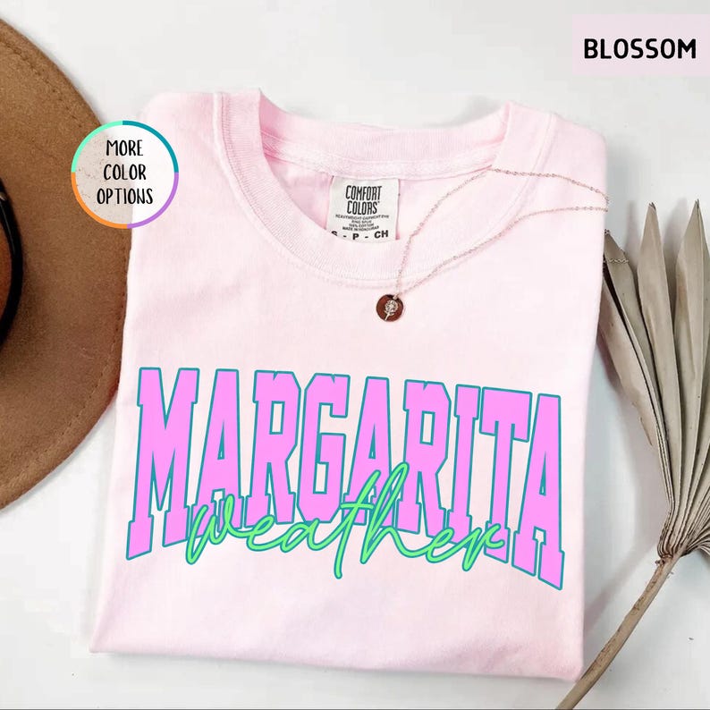May include: A light pink t-shirt with the words "MARGARITA WEATHER" in large, bold, pink and blue letters. The shirt is made of a soft material and has a round neckline. A gold necklace with a pendant is draped on the shirt.