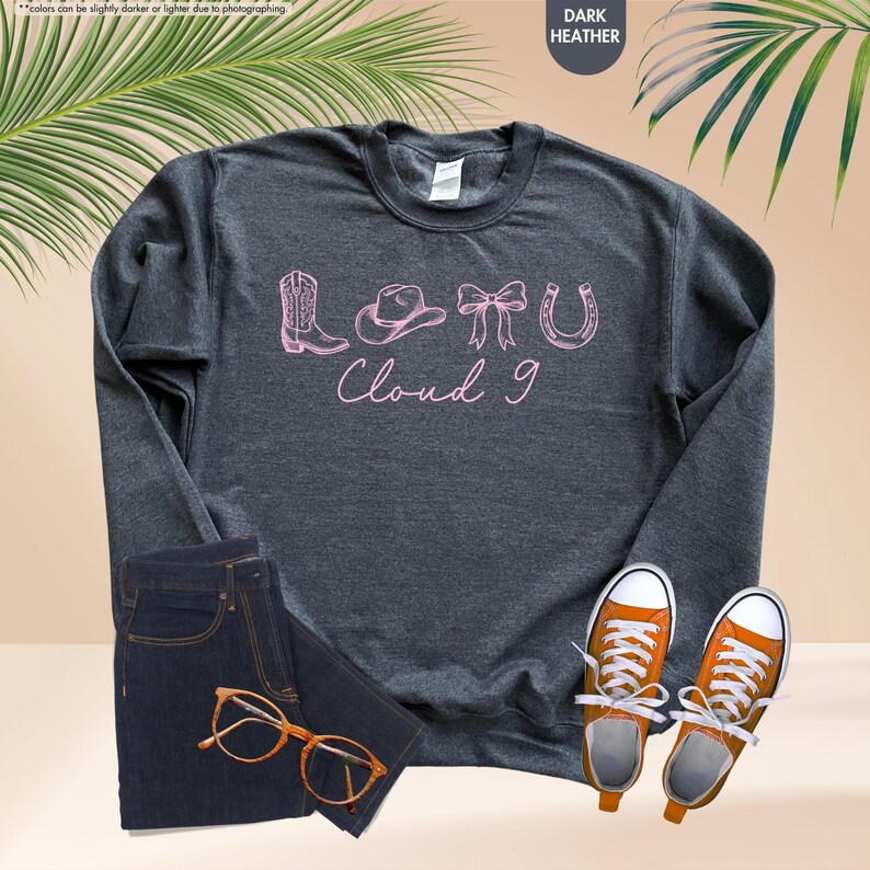 May include: Dark grey sweatshirt featuring a pink graphic of a cowboy boot, hat, bow, and horseshoe, with the text "Cloud 9". The image also shows dark wash jeans, orange trainers, and glasses.