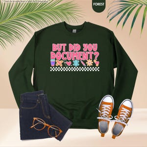 Puede incluir: Una sudadera verde oscuro con el texto "BUT DID YOU DOCUMENT?" en rosa y blanco. El dise&ntilde;o incluye gr&aacute;ficos de tem&aacute;tica m&eacute;dica y un borde a cuadros. La imagen tambi&eacute;n muestra unos vaqueros azules, zapatillas naranjas y gafas.