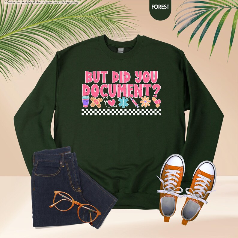 Puede incluir: Una sudadera verde oscuro con el texto "BUT DID YOU DOCUMENT?" en rosa y blanco. El dise&ntilde;o incluye gr&aacute;ficos de tem&aacute;tica m&eacute;dica y un borde a cuadros. La imagen tambi&eacute;n muestra unos vaqueros azules, zapatillas naranjas y gafas.