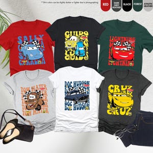以下が含まれることがあります： 様々な色の漫画の車のデザインが施されたTシャツのコレクション。赤、黒、グレーなど。サリー、グイド、ライトニング・マックィーンなどのキャラクターが描かれています。「Cars」の文字も確認できます。