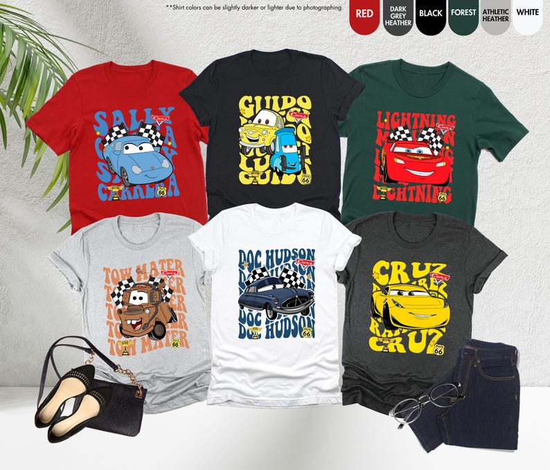 以下が含まれることがあります： 様々な色の漫画の車のデザインが施されたTシャツのコレクション。赤、黒、グレーなど。サリー、グイド、ライトニング・マックィーンなどのキャラクターが描かれています。「Cars」の文字も確認できます。