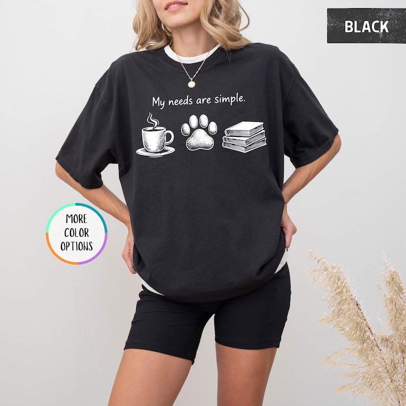 May include: Black t-shirt with the phrase "My needs are simple" and illustrations of a coffee cup, paw print, and books. The shirt has a relaxed fit and short sleeves. The words "More Color Options" are in a circle.