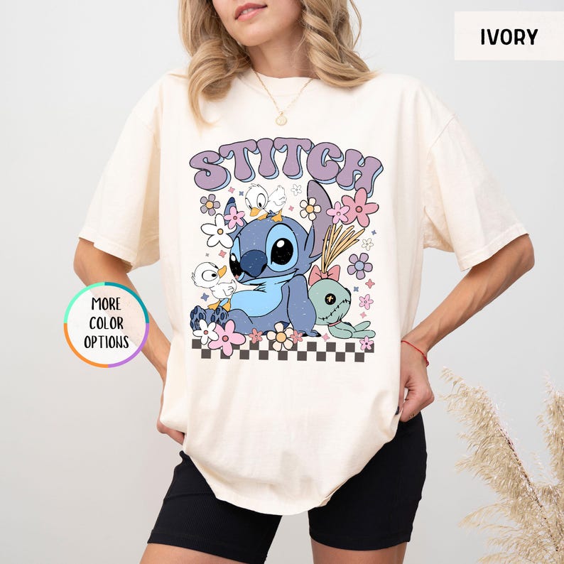 May include: Ivory-colored t-shirt featuring a graphic of Stitch with the word "STITCH" in a retro font. The design includes flowers, ducks, and a small plush toy. The shirt has short sleeves and a relaxed fit.