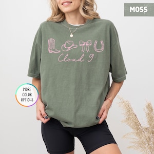 May include: A moss green t-shirt with a pink graphic featuring a cowboy boot, hat, bow, and horseshoe, with the text "Cloud 9". The shirt is a relaxed fit and has short sleeves.