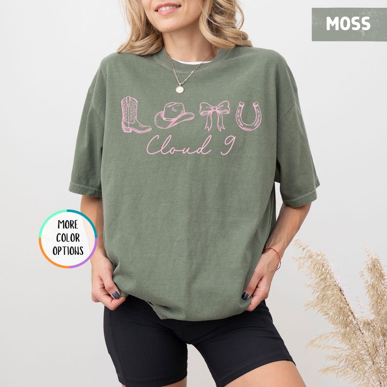 May include: A moss green t-shirt with a pink graphic featuring a cowboy boot, hat, bow, and horseshoe, with the text "Cloud 9". The shirt is a relaxed fit and has short sleeves.