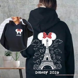 May include: A black hooded sweatshirt featuring a white graphic on the back of the Eiffel Tower, clouds, hearts, and a Minnie Mouse ear silhouette with a red polka dot bow. The text reads DISNEY 2026. The front shows a small Minnie ear logo and EMILY.