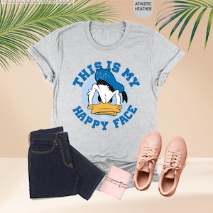 Op de afbeelding: Een lichtgrijs t-shirt met een afbeelding van Donald Duck en de tekst "THIS IS MY HAPPY FACE" in het blauw. De afbeelding bevat ook een donkerblauwe jeans, roze sneakers en een roze portemonnee.