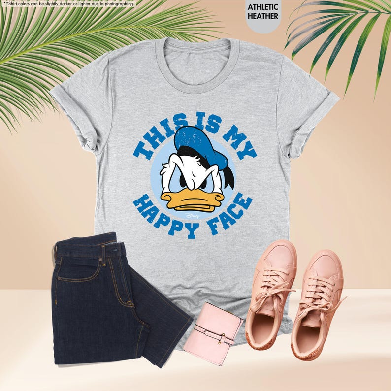 Op de afbeelding: Een lichtgrijs t-shirt met een afbeelding van Donald Duck en de tekst "THIS IS MY HAPPY FACE" in het blauw. De afbeelding bevat ook een donkerblauwe jeans, roze sneakers en een roze portemonnee.