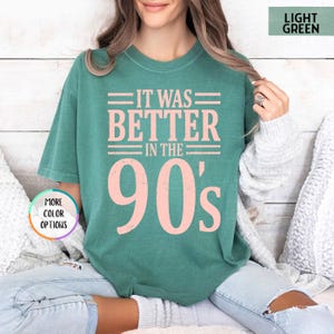 May include: A light green short-sleeve t-shirt featuring the text IT WAS BETTER IN THE 90's in a distressed pink font. The shirt includes a small circular graphic in the bottom left corner with the text MORE COLOR OPTIONS and a top right label.