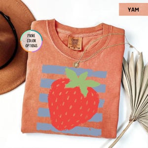 May include: A yam-colored short-sleeve t-shirt featuring a graphic of a red strawberry with a green stem centered over horizontal light blue stripes. The shirt has a crew neckline and a Comfort Colors brand label printed on the inside collar.