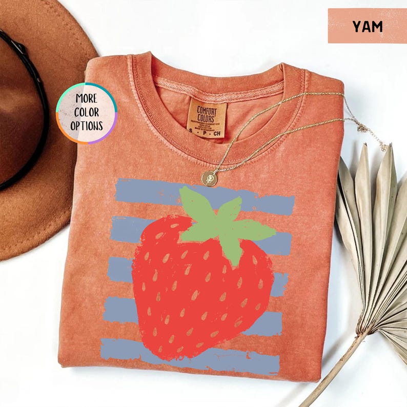 May include: A yam-colored short-sleeve t-shirt featuring a graphic of a red strawberry with a green stem centered over horizontal light blue stripes. The shirt has a crew neckline and a Comfort Colors brand label printed on the inside collar.