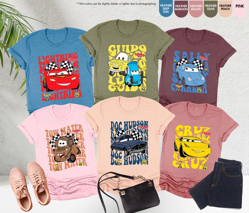 以下が含まれることがあります： アニメ映画「カーズ」のキャラクターが描かれた、様々な色のグラフィックTシャツ。デザインには、ライトニング・マックィーン、メーター、ドック・ハドソン、サリー、グイド、クルーズが含まれます。ピンクのスニーカー、黒いハンドバッグ、ジーンズと一緒に展示されています。