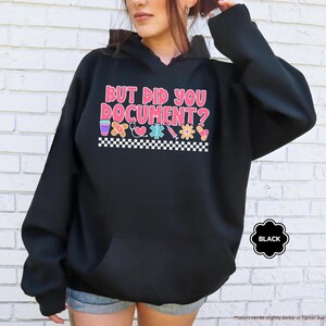 Puede incluir: Sudadera con capucha negra con el texto "BUT DID YOU DOCUMENT?" en rosa, rojo y azul. El dise&ntilde;o incluye iconos m&eacute;dicos y un borde a cuadros. Una etiqueta negra indica el color de la sudadera.