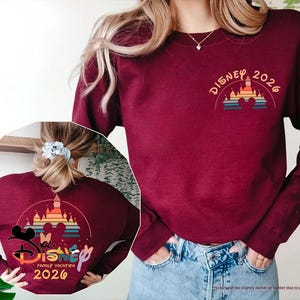 May include: Maroon sweatshirt with a Disney 2026 design. The design features a castle, Mickey and Minnie Mouse silhouettes, and the words "Disney Family Vacation 2026" in a colorful, arched layout. The sweatshirt has long sleeves.