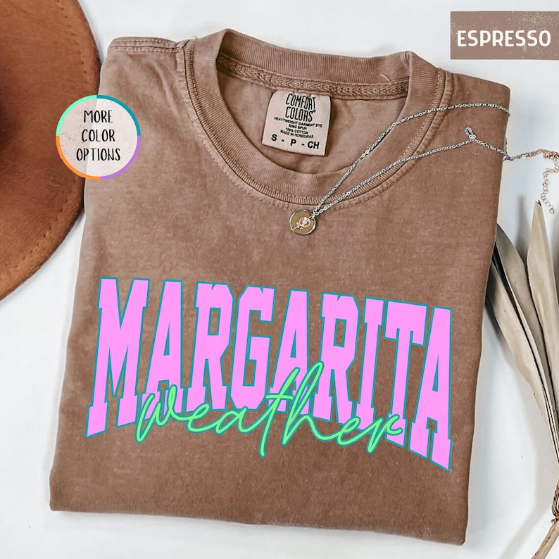May include: Espresso-colored t-shirt with the words "Margarita Weather" in pink and green. The shirt has a crew neck and short sleeves. A silver necklace with a pendant is draped on the shirt.