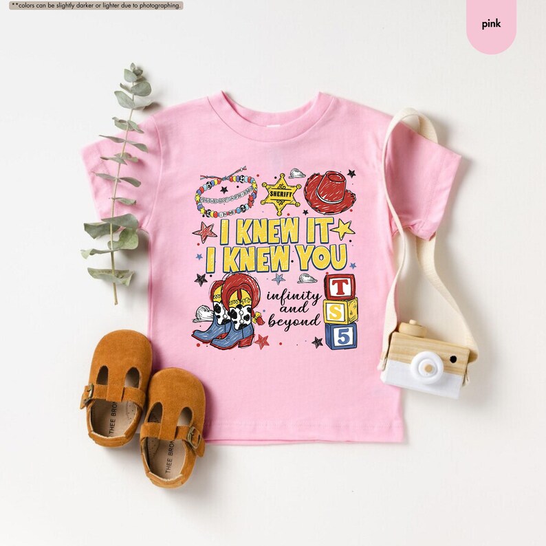 Peut inclure: Un t-shirt rose clair &agrave; manches courtes avec une impression graphique portant le texte I KNEW IT I KNEW YOU infinity and beyond. Le motif comprend un badge de sh&eacute;rif, un chapeau de cowboy rouge, des bottes et des cubes avec les lettres T, S, 5.