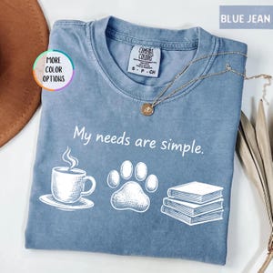 May include: Blue jean colored t-shirt with the phrase "My needs are simple" and illustrations of a coffee cup, paw print, and books. A gold necklace is draped across the shirt.
