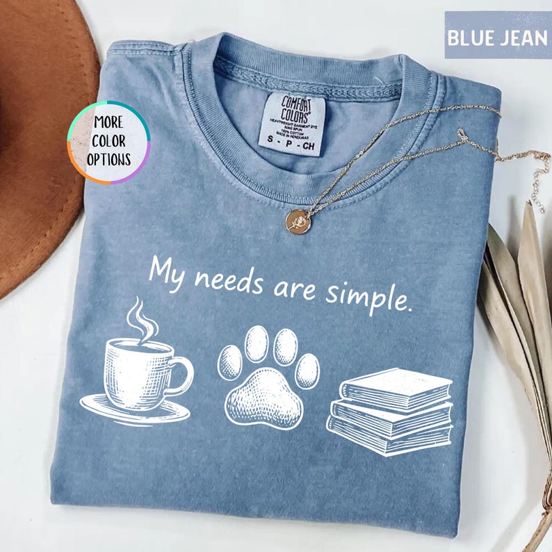 May include: Blue jean colored t-shirt with the phrase "My needs are simple" and illustrations of a coffee cup, paw print, and books. A gold necklace is draped across the shirt.