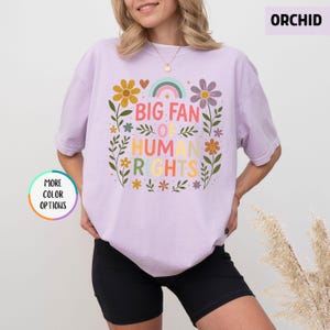 May include: Orchid-colored t-shirt with a floral design and the text "BIG FAN OF HUMAN RIGHTS" in colorful letters. The design includes a rainbow, flowers, and leaves. The shirt is a light purple color.