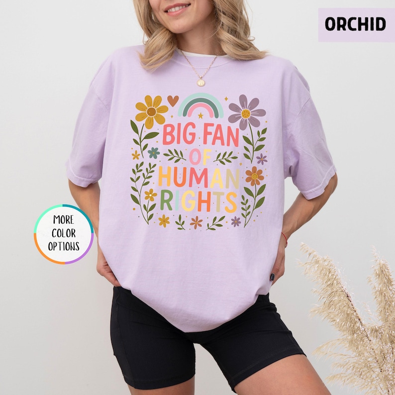 May include: Orchid-colored t-shirt with a floral design and the text "BIG FAN OF HUMAN RIGHTS" in colorful letters. The design includes a rainbow, flowers, and leaves. The shirt is a light purple color.