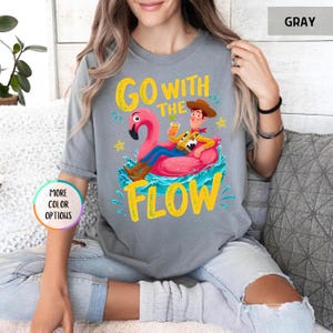 Puede incluir: Una camiseta gris de manga corta con un gr&aacute;fico de un vaquero de dibujos animados sentado en un flotador de flamenco rosa en agua azul. El texto "GO WITH THE FLOW" aparece en letras amarillas. La camiseta tiene cuello redondo.