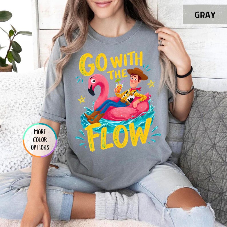 Puede incluir: Una camiseta gris de manga corta con un gr&aacute;fico de un vaquero de dibujos animados sentado en un flotador de flamenco rosa en agua azul. El texto "GO WITH THE FLOW" aparece en letras amarillas. La camiseta tiene cuello redondo.