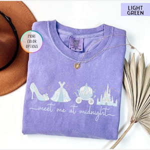 May include: A light purple Comfort Colors t-shirt with a graphic of a high heel, ball gown, carriage, and castle. The text "meet me at midnight" is written in a script font. A gold necklace is draped on the shirt.