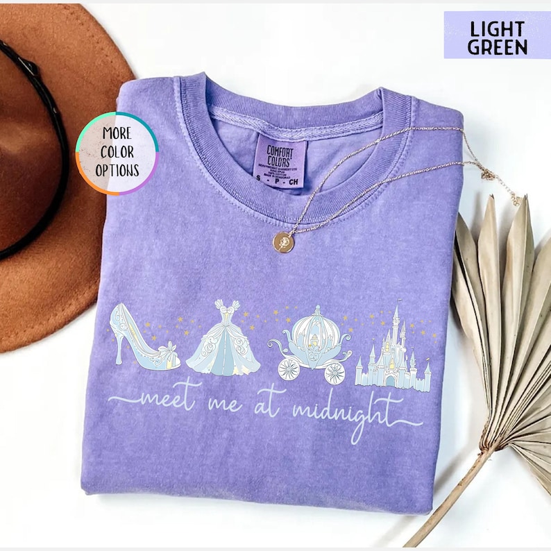 May include: A light purple Comfort Colors t-shirt with a graphic of a high heel, ball gown, carriage, and castle. The text "meet me at midnight" is written in a script font. A gold necklace is draped on the shirt.