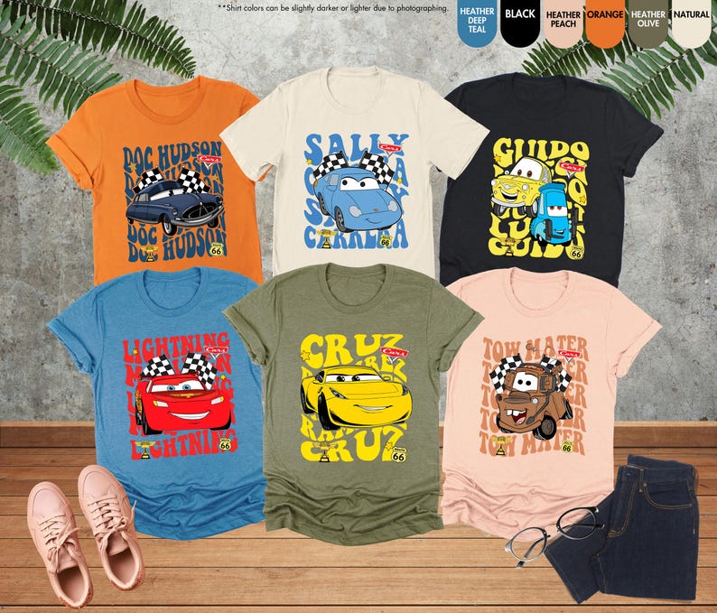 以下が含まれることがあります： 映画「カーズ」のキャラクターが描かれた、カラフルなTシャツのコレクション。オレンジ、青、黒、ピーチ、オリーブなど、さまざまな色があります。各シャツには、それぞれのキャラクターの名前と、チェッカーフラッグのデザインが表示されています。