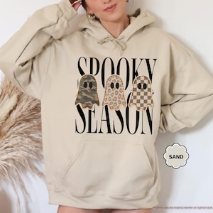 May include: A sand-colored hooded sweatshirt featuring the black text SPOOKY SEASON and three ghost illustrations. The ghosts have distinct patterns: one in camouflage, one in leopard print, and one in a brown and tan checkerboard pattern.