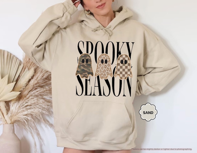 May include: A sand-colored hooded sweatshirt featuring the black text SPOOKY SEASON and three ghost illustrations. The ghosts have distinct patterns: one in camouflage, one in leopard print, and one in a brown and tan checkerboard pattern.