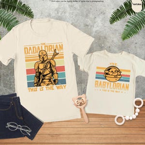 Puede incluir: Dos camisetas color crema con gr&aacute;ficos tem&aacute;ticos de Star Wars. Una camiseta dice "The Dadalorian" con una figura mandaloriana, y la otra dice "The Babylorian" con una imagen de Grogu. Ambas camisetas tienen la frase "This is the Way".