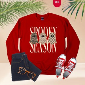 May include: A red long-sleeve crewneck sweatshirt featuring the text SPOOKY SEASON in white capital letters. Between the words are three ghost illustrations with different patterns: one in camouflage, one in leopard print, and one in a brown checkerboard.