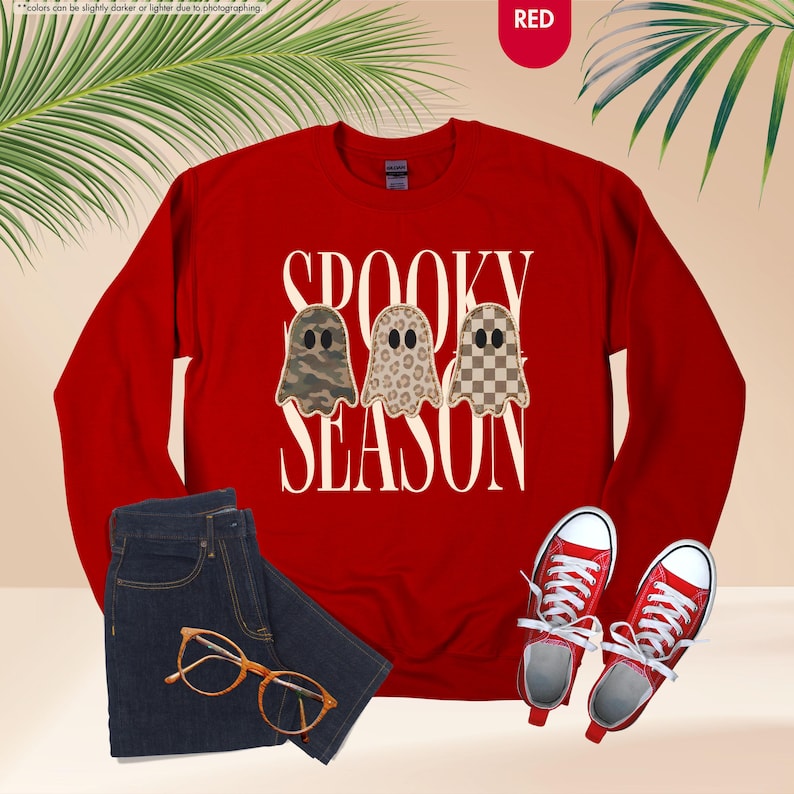 May include: A red long-sleeve crewneck sweatshirt featuring the text SPOOKY SEASON in white capital letters. Between the words are three ghost illustrations with different patterns: one in camouflage, one in leopard print, and one in a brown checkerboard.