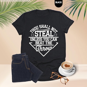 May include: Black t-shirt with the text "Thou Shall Not Steal Unless You Can Beat The Throw" in white. The shirt is paired with dark blue jeans, a cup of coffee with a heart design, and eyeglasses.