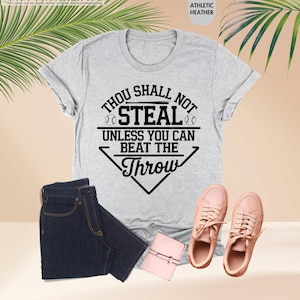 May include: A light gray t-shirt with the text "Thou Shall Not Steal Unless You Can Beat The Throw" in black. The shirt is paired with dark blue jeans, pink sneakers, and a small pink wallet. The shirt is athletic heather.