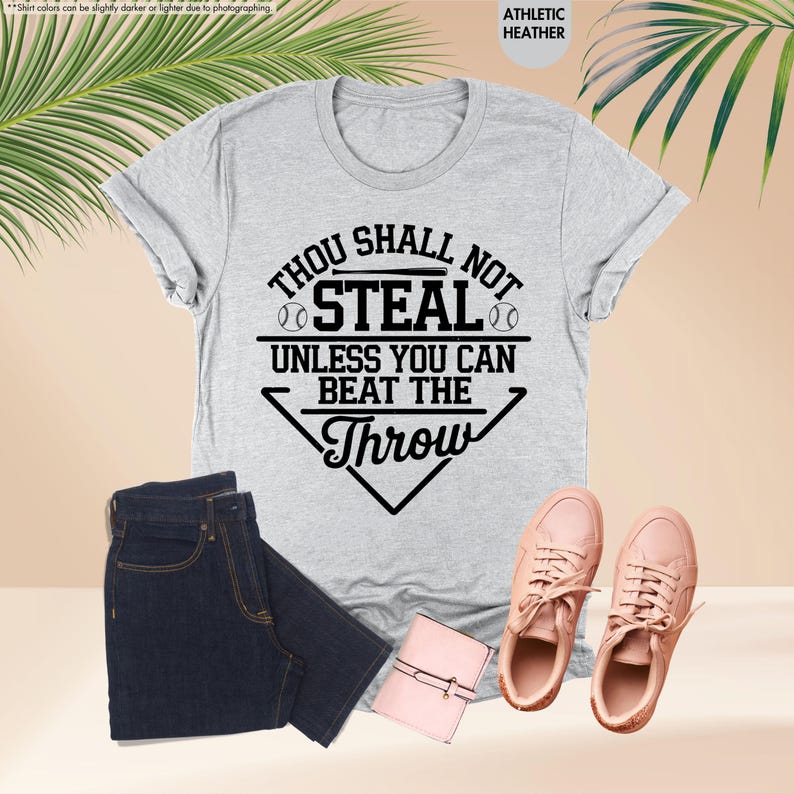 May include: A light gray t-shirt with the text "Thou Shall Not Steal Unless You Can Beat The Throw" in black. The shirt is paired with dark blue jeans, pink sneakers, and a small pink wallet. The shirt is athletic heather.