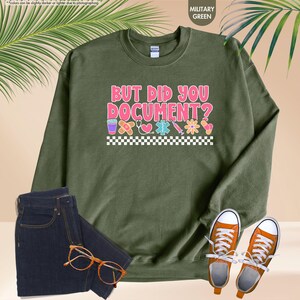 Puede incluir: Sudadera verde militar con el texto "BUT DID YOU DOCUMENT?" en rosa y blanco, con gr&aacute;ficos de tem&aacute;tica m&eacute;dica. La imagen tambi&eacute;n muestra vaqueros azul oscuro, zapatillas naranjas y gafas.