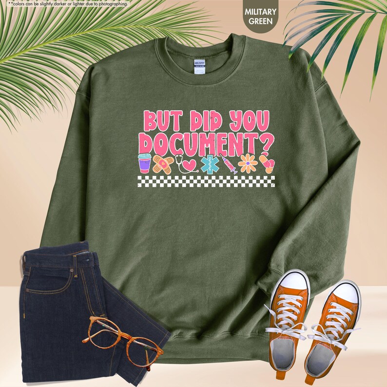 Puede incluir: Sudadera verde militar con el texto "BUT DID YOU DOCUMENT?" en rosa y blanco, con gr&aacute;ficos de tem&aacute;tica m&eacute;dica. La imagen tambi&eacute;n muestra vaqueros azul oscuro, zapatillas naranjas y gafas.