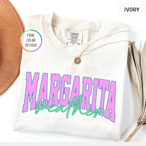 May include: Ivory-colored t-shirt with the words "Margarita Weather" in pink and green. The shirt has a crew neck and short sleeves. A gold necklace with a pendant is draped across the shirt.