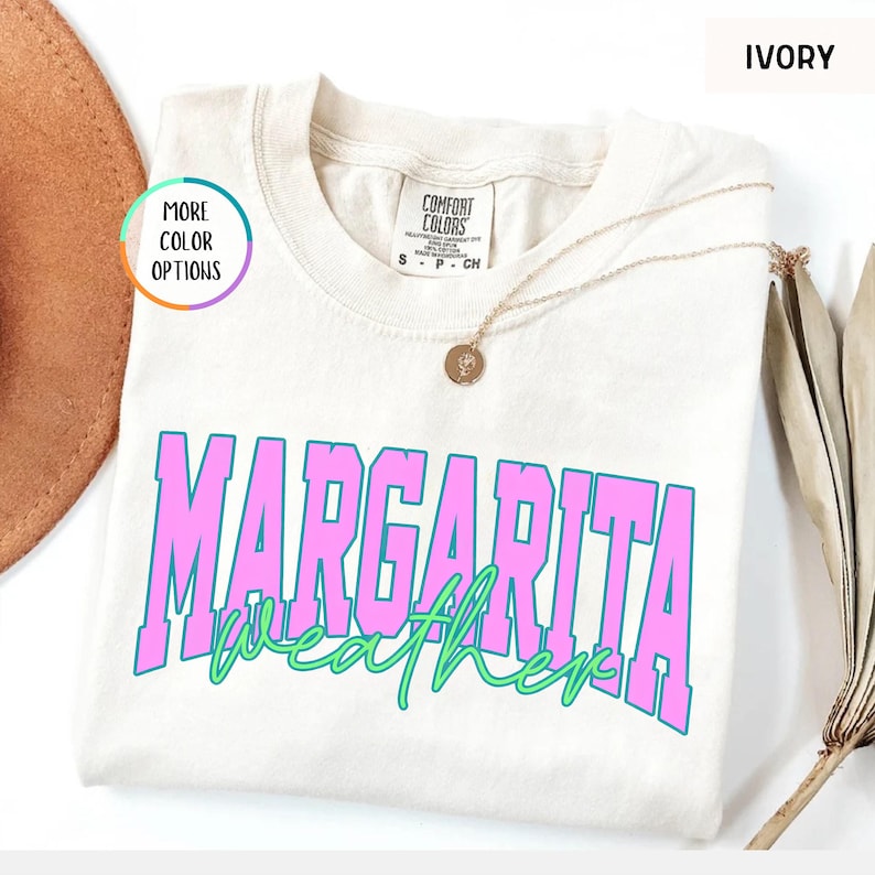 May include: Ivory-colored t-shirt with the words "Margarita Weather" in pink and green. The shirt has a crew neck and short sleeves. A gold necklace with a pendant is draped across the shirt.