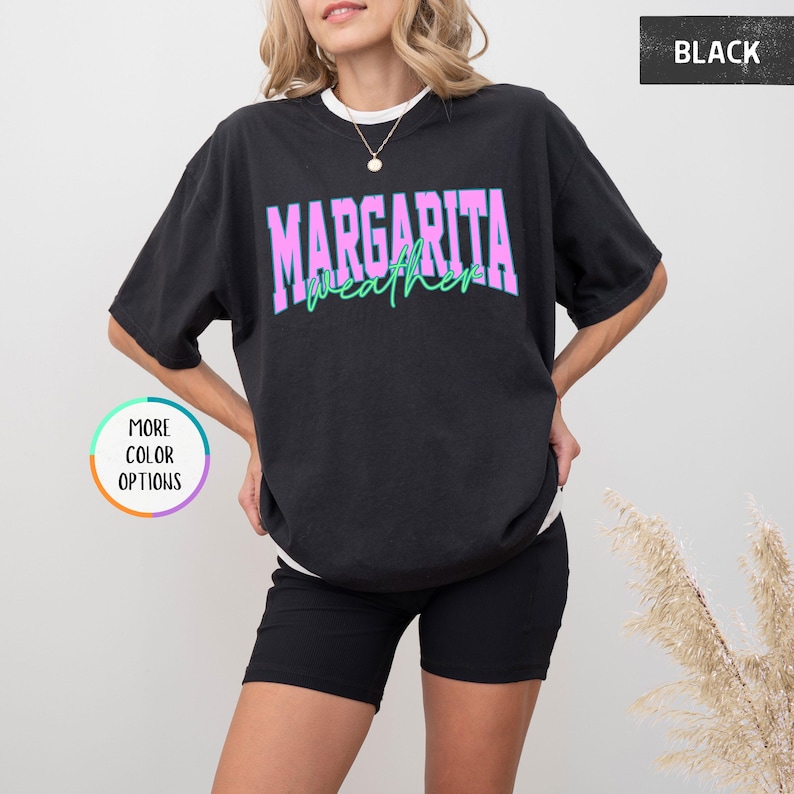 May include: Black t-shirt with the text "MARGARITA weather" in a pink and green design. The shirt has a relaxed fit and short sleeves. The image also shows a woman wearing the shirt.